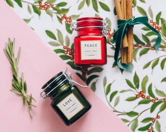 May include: Two small glass jars with lids, one red and one green, containing scented candles. The red candle is labelled "PEACE" with the scent "CINNAMON" and the green candle is labelled "LOVE" with the scent "WILDHONEY".
