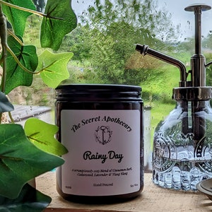 Op de afbeelding: Een zwarte kaars pot met een wit label waarop staat "The Secret Apothecary Rainy Day A scrumptiously cozy blend of Cinnamon Bark, Cedarwood, Lavender & Ylang Ylang Hand Poured Soy 8 oz".
