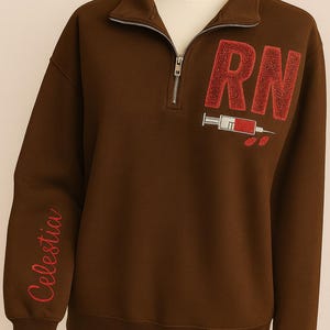 Könnte beinhalten: Ein braunes Sweatshirt mit Viertelreißverschluss, den Buchstaben "RN" in Rot, einer Spritzengrafik und dem Namen "Celestia" in roter Schrift auf dem Ärmel. Es hat einen gerippten Saum und Bündchen.