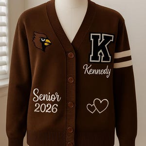 May include: Brown cardigan sweater with a V-neck and button closure. Features a cardinal bird patch, the word "Kennedy", the year "2026", and two hearts. White stripes on the sleeve.