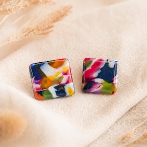 Könnte beinhalten: Zwei quadratische Ohrstecker mit einem farbenfrohen, abstrakten Design. Die Ohrringe weisen eine Mischung aus blauen, rosa, gelben und weißen Farbtönen auf. Sie sind vor einem neutralen Hintergrund platziert, der ihre lebendigen Farben und ihr einzigartiges Muster hervorhebt.