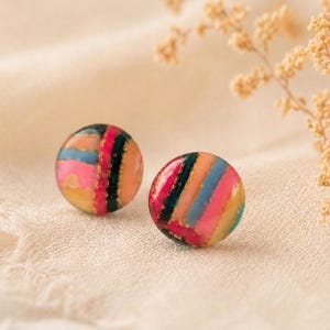 Könnte beinhalten: Zwei runde Ohrstecker mit einem farbenfrohen Streifendesign. Die Ohrringe weisen eine Mischung aus rosa, blauen, orangefarbenen und goldenen Streifen mit einem schwarzen vertikalen Streifen auf. Die Ohrringe sind vor einem neutralen Hintergrund platziert.