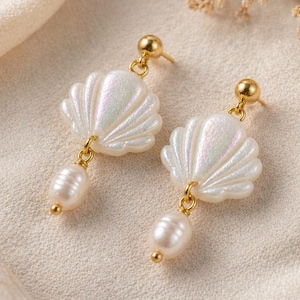 Könnte beinhalten: Ein Paar elegante Ohrringe mit Muscheldesign und schimmerndem Finish. Jeder Ohrring hat einen goldfarbenen Stecker und eine herabhängende Perle. Die Ohrringe sind auf einem cremefarbenen Stoff platziert.