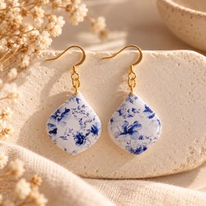 Könnte beinhalten: Ein Paar Ohrhänger mit tropfenförmigen weißen Anhängern, die mit einem blauen Blumenmuster versehen sind. Die Anhänger hängen an goldfarbenen Metallhaken. Die Oberfläche der Anhänger hat ein glattes, glänzendes Finish, das Licht reflektiert.