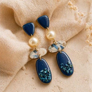 Könnte beinhalten: Ein Paar Ohrhänger in Marineblau und Gold. Jeder Ohrring hat eine dreieckige Spitze, eine große Perle, ein marmoriertes Oval und ein marineblaues Oval mit türkisfarbenen Akzenten. Die Ohrringe werden auf einer beigen Oberfläche präsentiert.