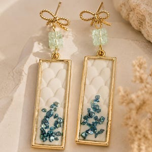 Könnte beinhalten: Ein Paar goldfarbene Ohrhänger mit rechteckigem Rahmen, weißem, texturiertem Schuppenmuster und eingebetteten blauen Metallsplittern. Jeder Ohrring ist mit einer kleinen hellgrünen quadratischen Perle und einer goldenen Schleife verziert.