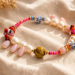 Könnte beinhalten: Ein Perlenarmband mit einer Mischung aus weißen Süßwasserperlen, leuchtend pinken Saatperlen und verschiedenen dekorativen Elementen, darunter eine blau-weiß gemusterte Perle, eine gesprenkelte senfgelbe Perle und hängende hellrosa Steinanhänger.