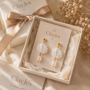 Könnte beinhalten: Ein Paar muschelförmige Ohrringe in Weiß mit goldfarbenen Steckern und hängenden weißen Perlen, präsentiert auf einer ClayJex-Markenkarte in einer weißen Geschenkbox mit Satinband. Die Karte zeigt den Text ClayJex und ein Blatt-Symbol.