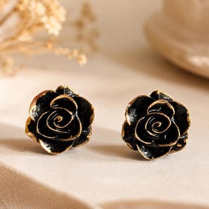 Könnte beinhalten: Ein Paar Ohrstecker in Form von blühenden Rosen. Jede Rose hat glänzend schwarze Blütenblätter mit metallisch goldfarbenen Rändern, die die geschwungenen Konturen der Blume betonen. Das Design ist symmetrisch und zeigt eine detaillierte Struktur.