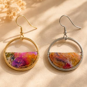 Könnte beinhalten: Ein Paar Ohrhänger mit kreisförmigen Metallrahmen. Ein Ohrring hat ein goldfarbenes Finish mit einem halbkreisförmigen Einsatz aus rosa, orangefarbenem und violettem Harz. Der andere Ohrring hat ein silberfarbenes Finish mit einem Einsatz aus orangefarbenem, violettem und weißem Harz.