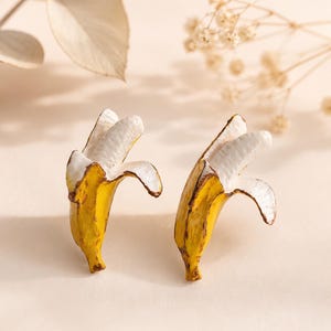 Könnte beinhalten: Ein Paar Ohrstecker in Form von teilweise geschälten Bananen. Jeder Ohrstecker zeigt eine leuchtend gelbe Schale mit braunen Details am Stiel und an der Spitze, die ein strukturiertes, weißes Fruchtfleisch im Inneren freilegt. Das Design ist realistisch.