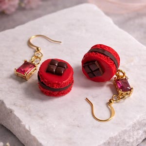 Könnte beinhalten: Goldfarbene Ohrringe mit roten Macaron-Anhängern und Schokoladendetails. Jeder Ohrring hat einen rechteckigen, roten Edelsteinanhänger. Die Ohrringe werden auf einer weißen, strukturierten Oberfläche präsentiert.