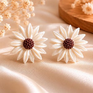 Könnte beinhalten: Ein Paar Ohrstecker in Form von Gänseblümchen mit glänzend weißen Blütenblättern und einer strukturierten dunkelbraunen Mitte. Das Blumendesign besteht aus mehreren spitzen Blütenblättern, die von einem runden, körnigen Kern ausgehen.