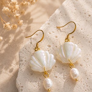 Könnte beinhalten: Ein Paar goldfarbene Ohrhänger mit einem weißen, irisierenden, muschelförmigen Anhänger, der über einer kleinen, ovalen weißen Perle hängt. Die Ohrringe sind mit einem einfachen Metallhaken zur Befestigung und einem feinen Goldglied versehen.