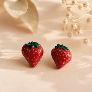 Könnte beinhalten: Ein Paar Ohrstecker in Form von Erdbeeren. Jeder Ohrstecker zeigt einen glänzend roten Fruchtkörper mit strukturierter Oberfläche und kleinen gelben Samen-Details, gekrönt von einem geformten grünen Blattstiel in dreidimensionaler Optik.