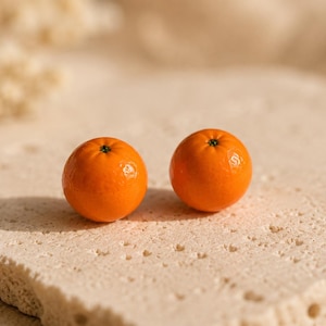 Könnte beinhalten: Zwei kleine, runde, leuchtend orangefarbene Fruchtkugeln mit glatter, glänzender Schale und einem winzigen dunkelgrünen Stielpunkt an der Oberseite. Diese miniaturartigen, zitrusähnlichen Objekte sind nebeneinander platziert und zeigen eine kräftige Farbe.