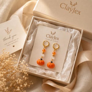 Könnte beinhalten: Ein Paar goldfarbene Creolen mit orangefarbenen Kürbisanhängern, klaren facettierten Perlen und kleinen orangefarbenen Perlen. Die Ohrringe sind auf einer ClayJex-Karte in einer Geschenkbox mit Dankeskarte und Seidenpapier präsentiert.