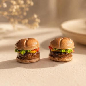 Könnte beinhalten: Zwei Miniatur-Hamburger-Figuren mit einem beigen Brötchen, einem dunkelbraunen Patty, einer Scheibe gelbem Käse und hellgrünem Salat. Die Burger haben eine glänzende Oberfläche und realistische Texturen mit einer kreuzförmigen Vertiefung im Brötchen.