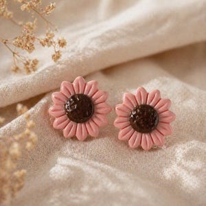 Könnte beinhalten: Ein Paar rosa Sonnenblumenohrringe mit dunkelbraunen Zentren. Die Ohrringe sind vor einem neutralfarbenen Stoffhintergrund platziert, mit getrockneten floralen Akzenten. Ein floraler Schmuck.