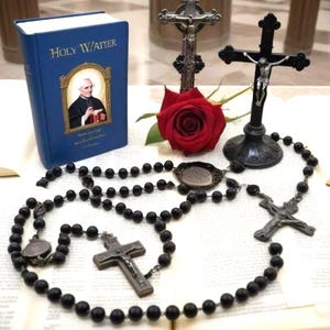 May include: A still life composition with a blue book labelled "Holy Watter", a red rose, rosary beads, and crucifixes. The rosary beads are black, and the crucifixes are silver and black. The book is open on a white surface.
