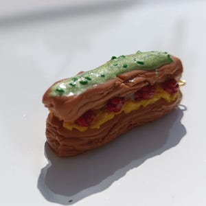 Könnte beinhalten: Ein detailliertes Miniatur-Eclair mit braunem Gebäckboden, gelber Creme, roten Beeren und grüner Glasur. Dieses handgefertigte Dessert ist eine Miniatur-Replik.