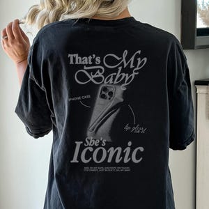 May include: Black oversized t-shirt with the text "That's My Baby" and "She's Iconic" in a stylized font. The design includes an illustration of a phone case and the words "lip gloss on it".
