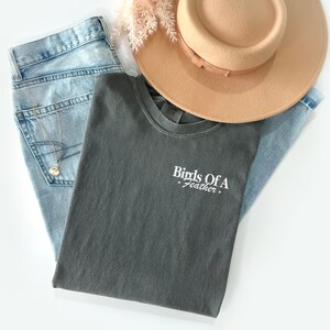 Peut inclure: Un t-shirt gris fonc&eacute; avec le texte "Birds Of A Feather" brod&eacute; en blanc sur le devant. Le t-shirt est pli&eacute; et pos&eacute; sur un jean bleu clair et un fedora brun.