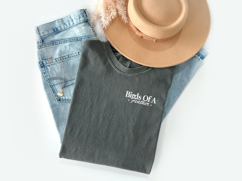 Peut inclure: Un t-shirt gris fonc&eacute; avec le texte "Birds Of A Feather" brod&eacute; en blanc sur le devant. Le t-shirt est pli&eacute; et pos&eacute; sur un jean bleu clair et un fedora brun.
