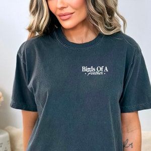 Peut inclure: Un t-shirt gris fonc&eacute; avec le texte "Birds Of A Feather" imprim&eacute; &agrave; l'avant en blanc.