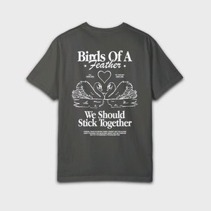 Peut inclure: Un t-shirt gris fonc&eacute; avec un imprim&eacute; graphique blanc de deux cygnes se faisant face avec un c&oelig;ur au milieu. Le texte "Birds of a Feather" est au-dessus des cygnes et le texte "We Should Stick Together" est en dessous des cygnes.