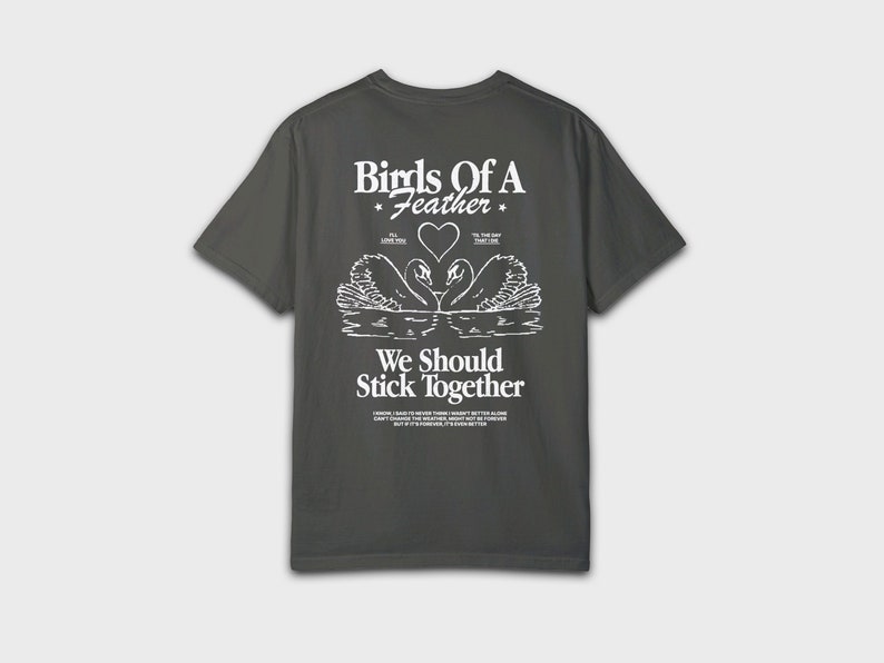Peut inclure: Un t-shirt gris fonc&eacute; avec un imprim&eacute; graphique blanc de deux cygnes se faisant face avec un c&oelig;ur au milieu. Le texte "Birds of a Feather" est au-dessus des cygnes et le texte "We Should Stick Together" est en dessous des cygnes.