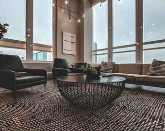 May include: A modern living room with large windows, a brown leather armchair, a black leather armchair, a brown sofa with pillows, and a black metal coffee table with a potted plant on top. The room has a large rug with a geometric pattern.