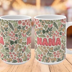 K&ouml;nnte beinhalten: Wei&szlig;e Keramiktasse mit einem Blumenmuster und dem Wort "NANA" in rosa Buchstaben. Die Tasse hat einen Henkel auf der rechten Seite.