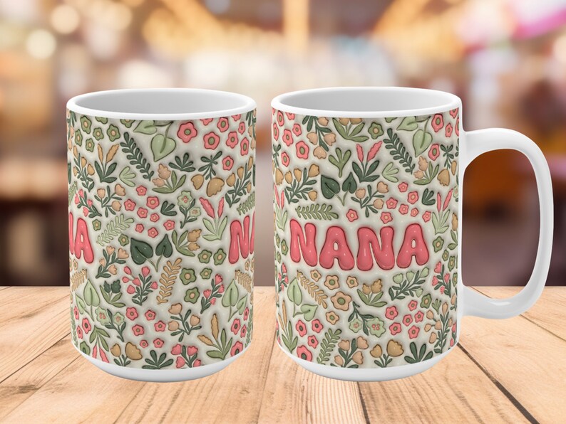 K&ouml;nnte beinhalten: Wei&szlig;e Keramiktasse mit einem Blumenmuster und dem Wort "NANA" in rosa Buchstaben. Die Tasse hat einen Henkel auf der rechten Seite.
