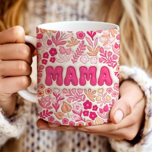 May include: White ceramic mug with a floral pattern in shades of pink, peach, and cream. The word "MAMA" is displayed in large, bubble-style pink letters. The mug has a white handle.