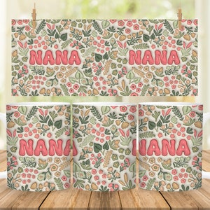 K&ouml;nnte beinhalten: Drei wei&szlig;e Keramikbecher mit einem Blumenmuster und dem Wort "NANA" in rosa Buchstaben. Die Becher stehen auf einer Holzoberfl&auml;che.