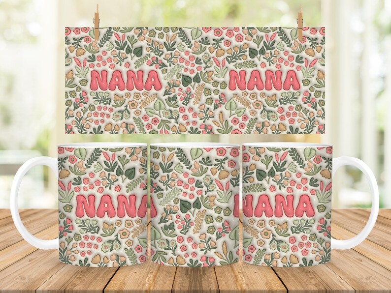 K&ouml;nnte beinhalten: Drei wei&szlig;e Keramikbecher mit einem Blumenmuster und dem Wort "NANA" in rosa Buchstaben. Die Becher stehen auf einer Holzoberfl&auml;che.