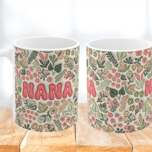 K&ouml;nnte beinhalten: Wei&szlig;e Keramiktasse mit einem Blumenmuster in Gr&uuml;n-, Rosa- und Braunt&ouml;nen. Das Wort "NANA" ist in rosa Buchstaben auf der Tasse geschrieben.