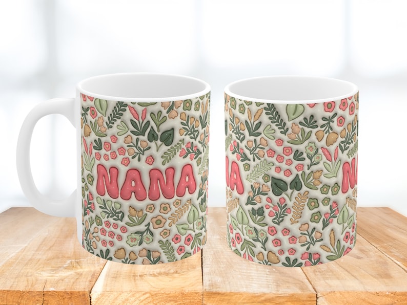 K&ouml;nnte beinhalten: Wei&szlig;e Keramiktasse mit einem Blumenmuster in Gr&uuml;n-, Rosa- und Braunt&ouml;nen. Das Wort "NANA" ist in rosa Buchstaben auf der Tasse geschrieben.