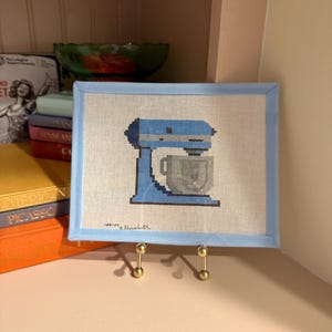 May include: A framed needlepoint featuring a blue stand mixer with a gray bowl. The artwork is set against a white background and framed with a light blue border. The piece is displayed on a gold-colored stand.