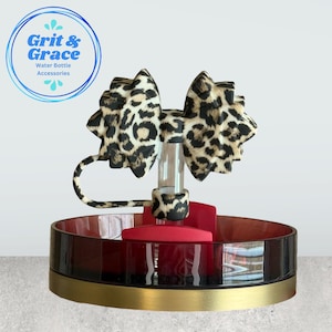 Op de afbeelding: Een waterflesaccessoire met een doorzichtig rietje en een luipaardprint strik. Het accessoire staat op een rood-zwarte basis met een gouden rand. De tekst "Grit & Grace Water Bottle Accessories" staat in een blauwe cirkel.