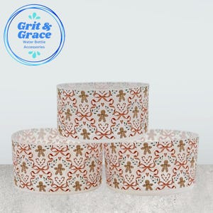 Può includere: Tre accessori per bottiglie d'acqua con un motivo festivo a omino di pan di zenzero. Il design include omini di pan di zenzero, fiocchi rossi, cuori e rametti verdi su sfondo bianco. Gli accessori sono cilindrici e riportano la scritta "Grit & Grace Water Bottle Accessories".