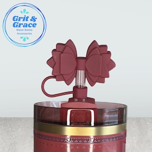 May include: A maroon water bottle accessory with a bow-shaped top. The water bottle has a maroon lid with a gold band and the text "Grit & Grace Water Bottle Accessories" in a blue circle.