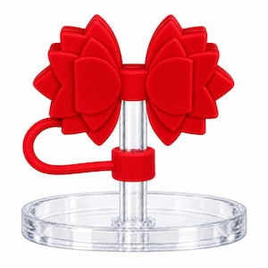 Puede incluir: Un adorno de silicona roja con forma de lazo unido a una pajita de plástico transparente y una base circular transparente. El lazo presenta un diseño en capas similar a pétalos con un bucle rojo flexible que se extiende desde el centro.
