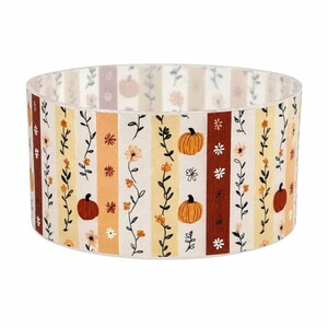 May include: A cylindrical lampshade featuring a repeating vertical striped pattern. The design includes alternating bands of cream, yellow, and rust-brown, decorated with illustrations of orange pumpkins, small white flowers, and delicate black leafy vines.