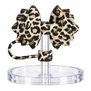 Puede incluir: Un accesorio en forma de lazo con estampado de leopardo en negro y beige, unido a un soporte de plástico transparente con base circular. El lazo tiene un diseño estructurado de varias capas con un pequeño bucle que se extiende hacia un poste.