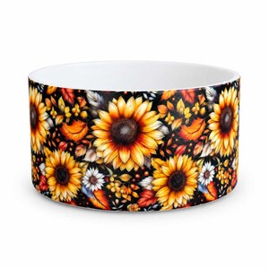 May include: A cylindrical pet bowl with a white interior and a black exterior featuring a vibrant pattern of yellow sunflowers, orange autumn leaves, small white flowers, and scattered berries across the entire outer surface.