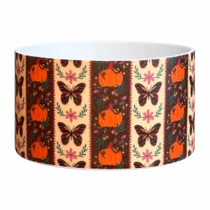 May include: A cylindrical ceramic bowl featuring a repeating pattern of orange pumpkins, brown butterflies, and pink flowers on alternating cream and dark brown vertical stripes. The interior of the bowl is solid white with a smooth finish.