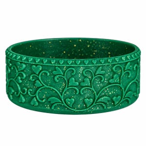 May include: A circular green bangle bracelet featuring a raised relief pattern of swirling vines and heart shapes. The surface of the bracelet is embedded with fine gold-colored glitter particles throughout the dark green material.