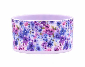 May include: A cylindrical bowl featuring a light lavender base color and an all-over floral pattern. The design consists of small, densely packed flowers in various shades of purple, blue, and pink, creating a vibrant and colorful botanical display.